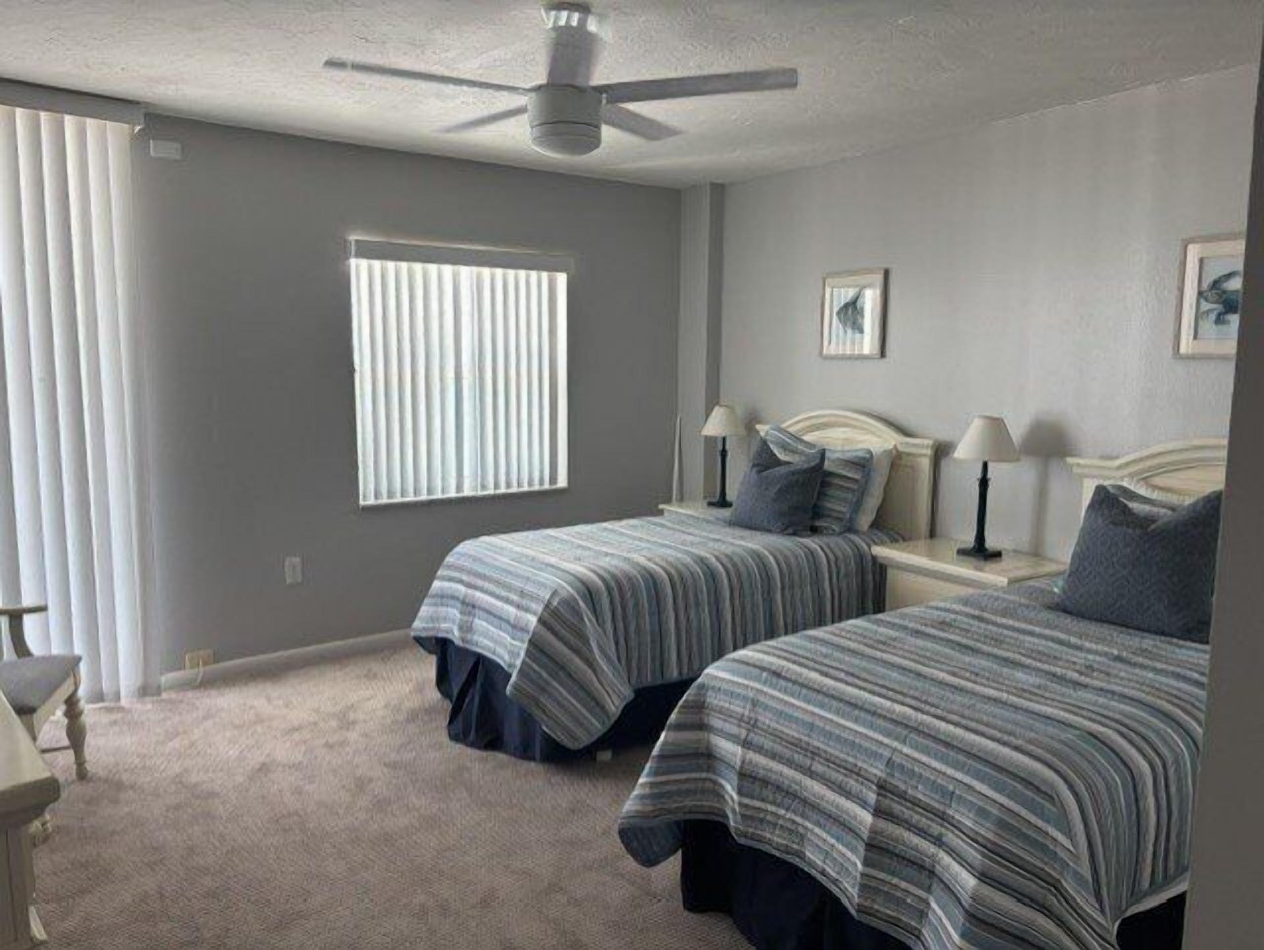 A bedroom with two beds, a ceiling fan, a window with blinds, and a framed picture on the wall.