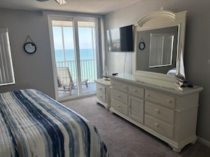 A bedroom with a bed, a dresser, a TV, and a balcony with a chair. - Crystal Sands Tower Unit 1202 - Beachfront (SARASOTA)