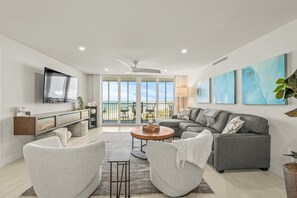 Una sala de estar moderna con un sofá gris, dos sillones, una mesita de centro y un televisor montado en la pared. Hay un balcón con vistas al océano.