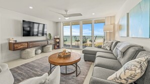 A spacious living room with a large flat-screen TV, a grey sectional sofa, a wooden coffee table, and a balcony with a view of the beach. - Crystal Sands Tower Unit 909 - Beachfront (Sarasota)