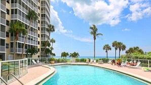 A swimming pool with lounge chairs and palm trees, surrounded by a high-rise building with balconies. - Crystal Sands Tower Unit 909 - Beachfront (Sarasota)