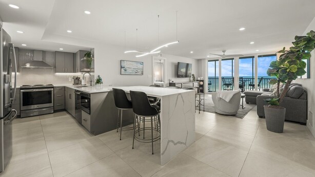 A modern kitchen with a central island, bar stools, and a dining area with a view of the ocean. - Crystal Sands Tower Unit 909 - Beachfront (Sarasota)