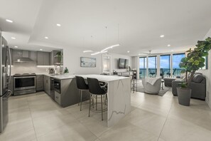 A modern kitchen with a central island, bar stools, and a dining area with a view of the ocean. - Crystal Sands Tower Unit 909 - Beachfront (Sarasota)
