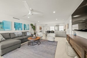 Una sala moderna con un sofá gris, una mesita de centro y un sillón blanco. Se ve una cocina al fondo con un refrigerador de acero inoxidable y un ventanal grande.