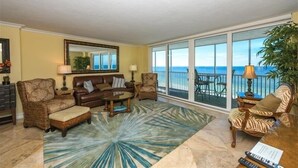 A living room with a large window overlooking the ocean, furnished with a sofa, chairs, and a coffee table. - Crystal Sands Tower Unit 703 - Beachfront (Sarasota)