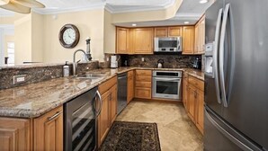 A kitchen with granite countertops, stainless steel appliances, wooden cabinets, and a clock on the wall. - Crystal Sands Tower Unit 703 - Beachfront (Sarasota)