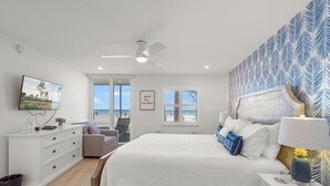 Een slaapkamer met een bed, een dressoir, een stoel, een televisie en een raam met uitzicht op het strand.