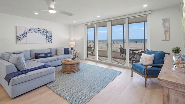 Een ruime woonkamer met een grijze bank, een blauwe fauteuil en een rieten salontafel. Grote glazen deuren leiden naar een balkon met uitzicht op het strand.