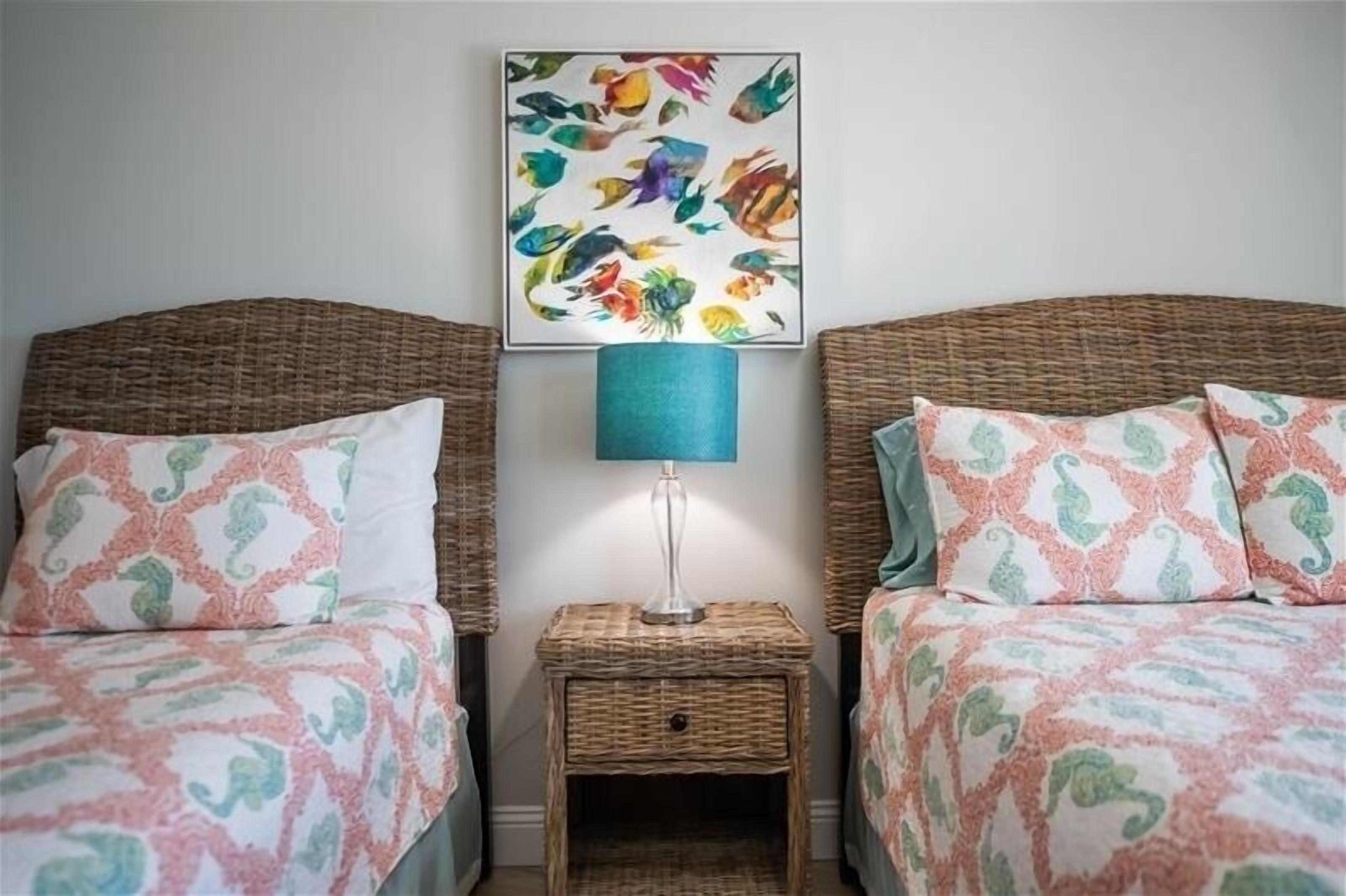 Une chambre avec deux lits, une table de chevet avec une lampe et un tableau coloré représentant des poissons accroché au mur.