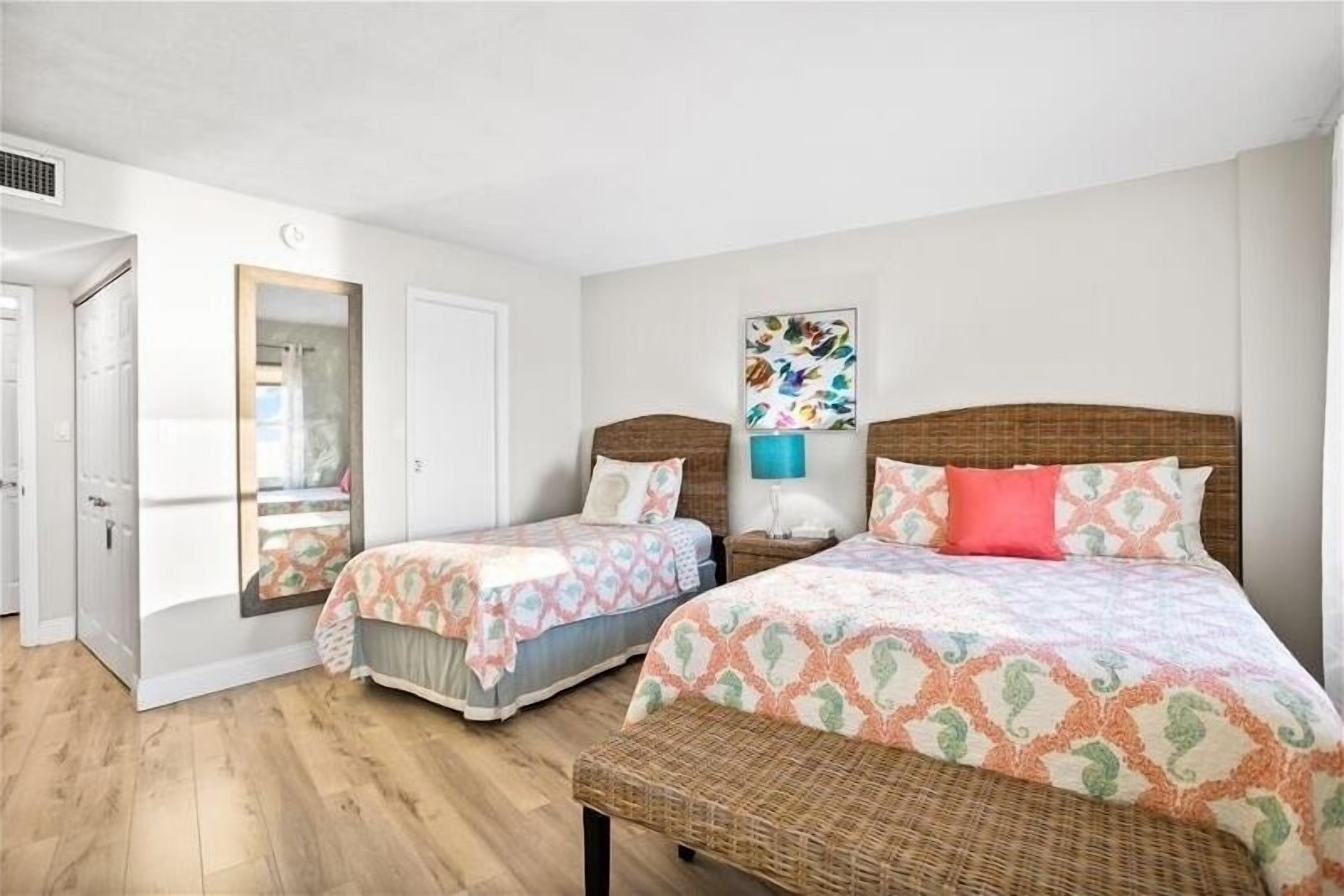 A bedroom with two beds, a wooden floor, a mirror, a wall art piece, and a bench.
