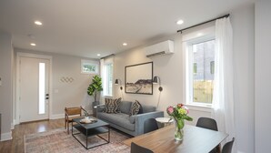 Una sala moderna con un sofá, una mesa de centro y una mesa de comedor. Hay una ventana con cortinas, un cuadro en la pared y un equipo de aire acondicionado.