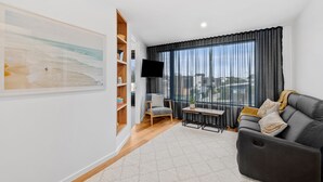 A modern living room with a grey sofa, a wooden bookshelf, a flat-screen TV, and a large window with curtains. - Apartment Studio 6: In The Heart of Torquay (Torquay)