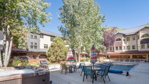Ein Whirlpool, ein Außenpool und Terrassenmöbel auf einer Holzterrasse, im Hintergrund Bäume und Gebäude.