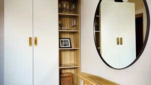 A room with a herringbone wooden floor, a white wardrobe with brass handles, a wooden shelf unit with glassware, a round mirror, and a small wooden table.