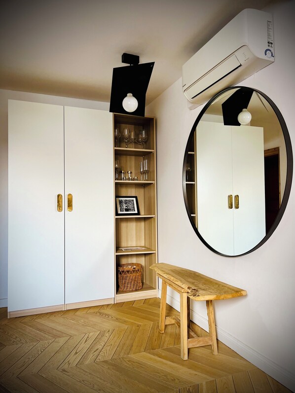 Een kamer met een visgraat parketvloer, een witte garderobe met messing handgrepen, een houten plankenwand met glaswerk, een ronde spiegel en een klein houten tafeltje.