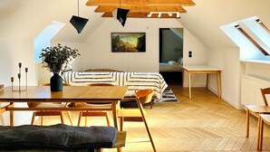 A spacious attic bedroom with a sloped ceiling, wooden beams, a bed with a striped bedspread, a dining table, and a vase with flowers.