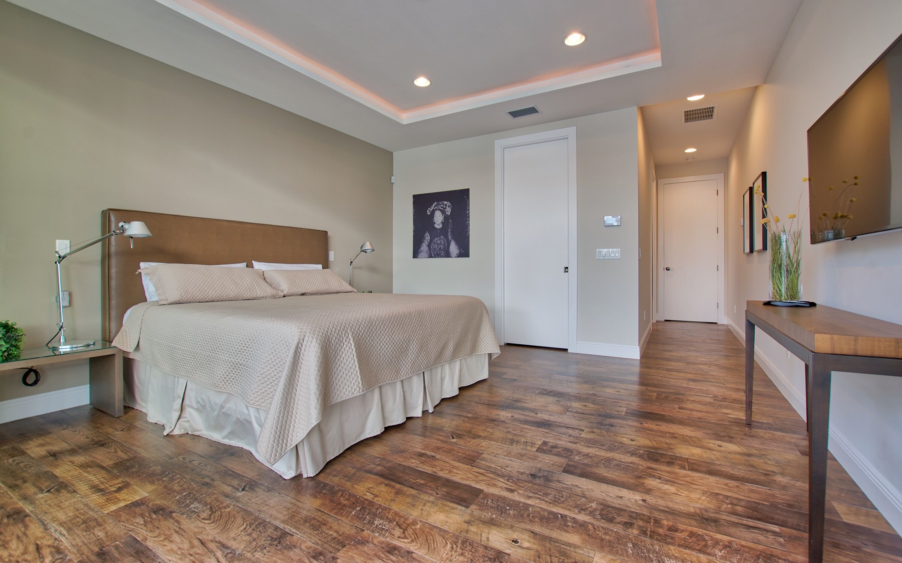 Een slaapkamer met een groot bed, bedlampjes, een nachtkastje, een houten vloer, een deur en een gang die naar een andere kamer leidt.