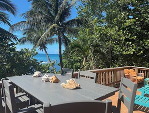 A table with chairs and a wooden deck overlooking a beach with palm trees and the ocean.