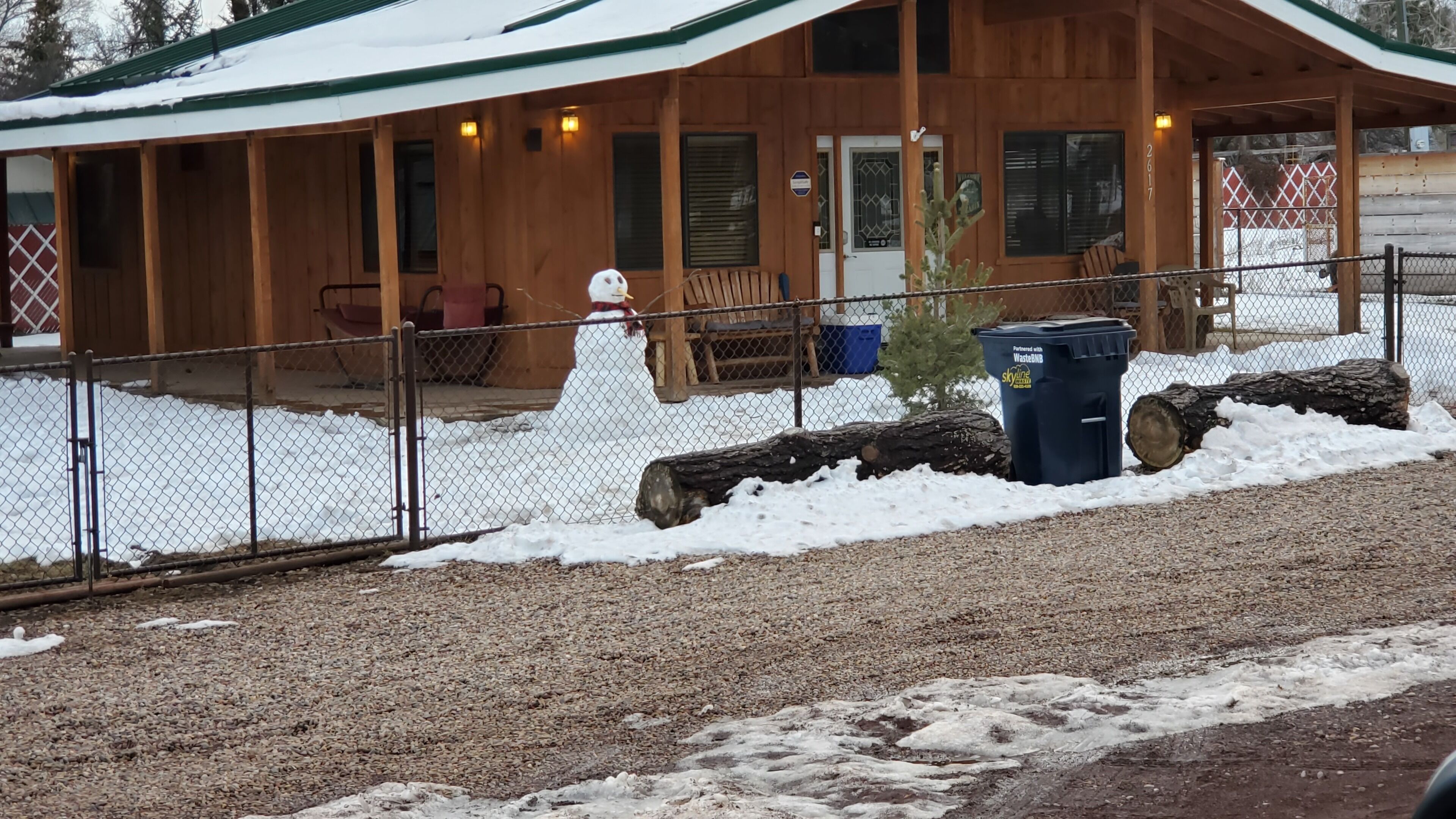 A wooden lodge with a covered porch, a fenced area, and a snow-covered ground.
