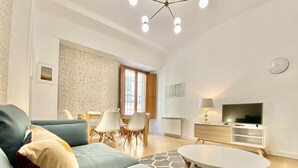 A modern living room with a sofa, a coffee table, and a TV. There is a dining area with chairs and a table. The room has a large window and a patterned rug. - Opera, Duplex Apartment – Three-bedroom Duplex Apartment (Madrid)