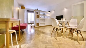 Uma sala de estar ampla com piso em padrão herringbone, uma mesa de jantar com cadeiras brancas, um sofá vermelho e estantes embutidas com diversos objetos.