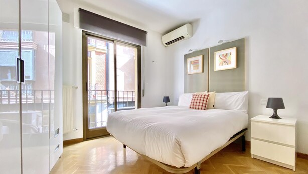 Quarto de hotel com cama, mesa de cabeceira com abajur, guarda-roupa e porta de vidro deslizante para a varanda.