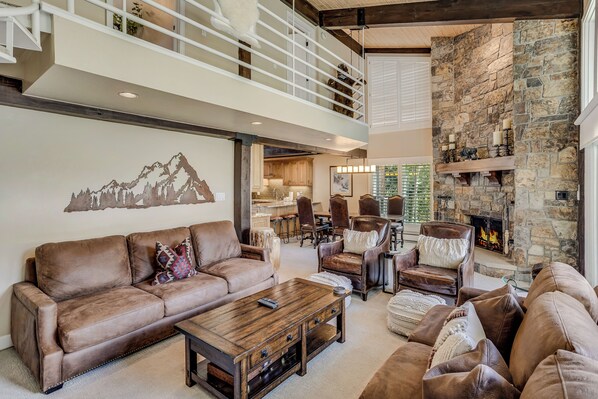 A spacious living area with a high ceiling, a stone fireplace, and a view of the kitchen.