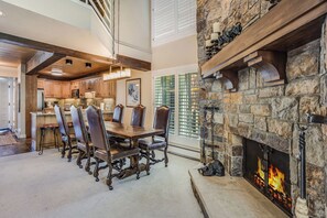 A spacious dining area with a stone fireplace, wooden beams, and a view of the kitchen.