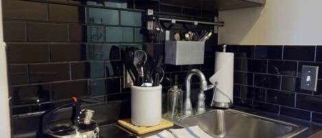 Une cuisine avec un dosseret de carreaux noirs, des appareils en acier inoxydable, une machine à café et des ustensiles rangés dans un porte-ustensiles.