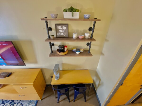 A compact room with a wooden desk, two chairs, a small shelf with decorative items, and a sign that reads "ENJOY YOUR STAY."