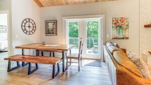 A dining area with a wooden table and benches, a clock, a painting, and a sofa with a view to the outdoors. - Deck, Fire Pit & Fireplace: Murphy Forest Retreat (Murphy)