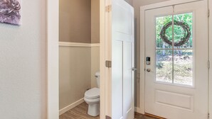 A bathroom with a toilet, a door with a wreath, and a window with a view of greenery. - Deck, Fire Pit & Fireplace: Murphy Forest Retreat (Murphy)