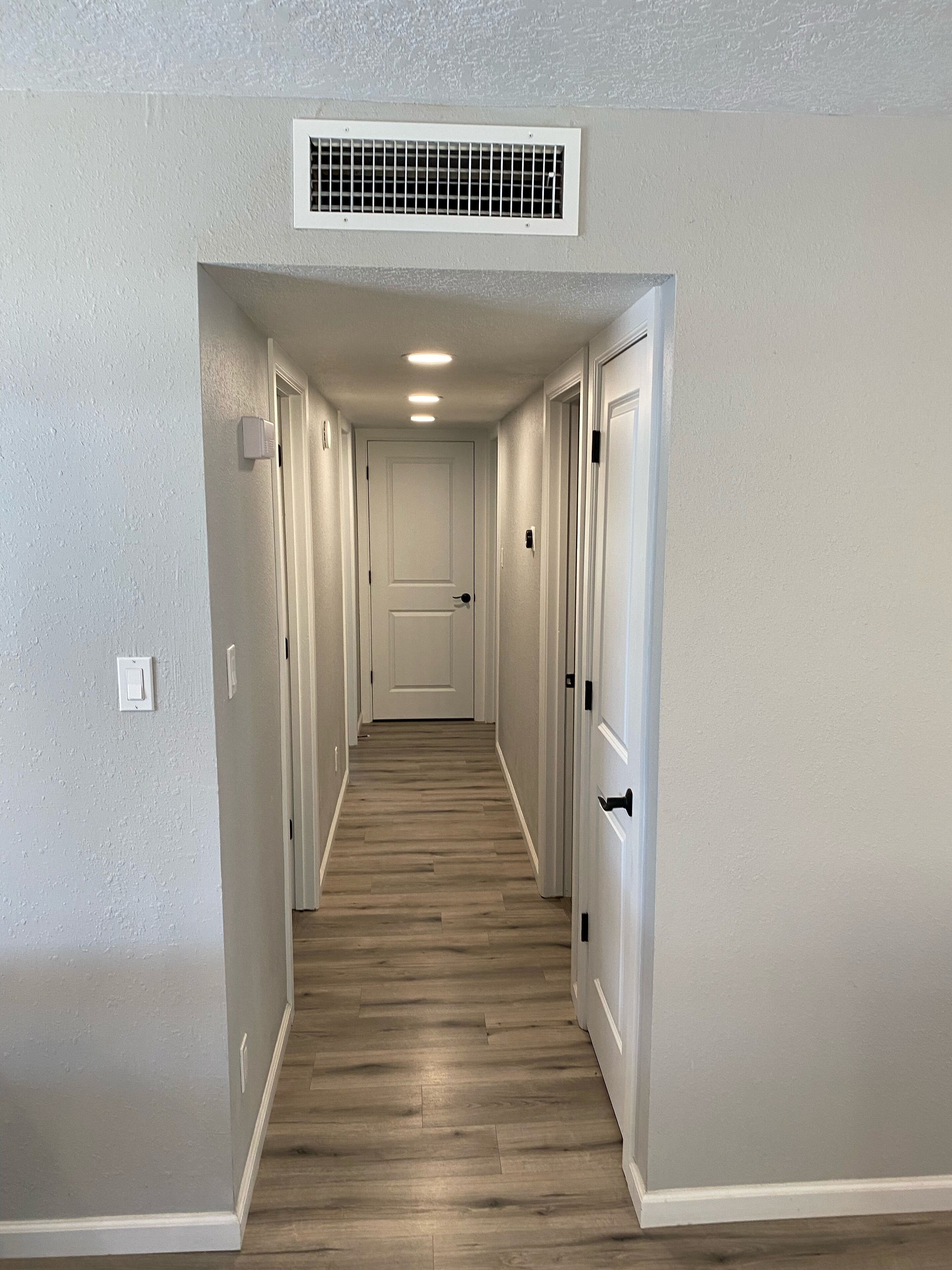 A hallway with a wooden floor, white walls, and a ceiling vent.