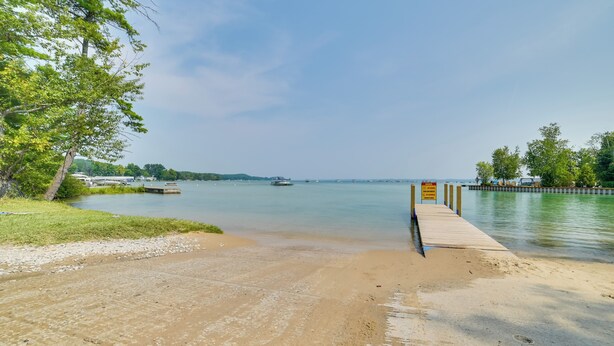 Een zanderig strand met een houten pier die zich uitstrekt over kalm water, omringd door bomen en verre bootjes.