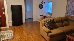Un salon avec un canapé beige, un réfrigérateur noir, un plancher en bois, un coin repas avec une table et des chaises, et un tapis à motifs.