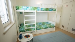 Een stapelbed met beddengoed met bladmotieven, een tafeltje met een vaas en een tijdschrift, en een raam met gordijnen.