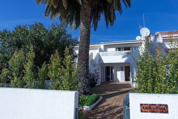 A white building with a paved entrance, surrounded by greenery and a clear blue sky.