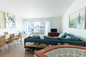 A modern living room with a sofa, a dining table, and chairs. There is a balcony with a view of the landscape.