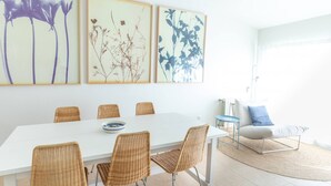 Een eethoek met een witte tafel, rieten stoelen en ingelijste botanische prints aan de muur.
