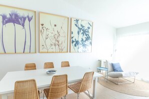 A dining area with a white table, wicker chairs, and framed botanical prints on the wall.