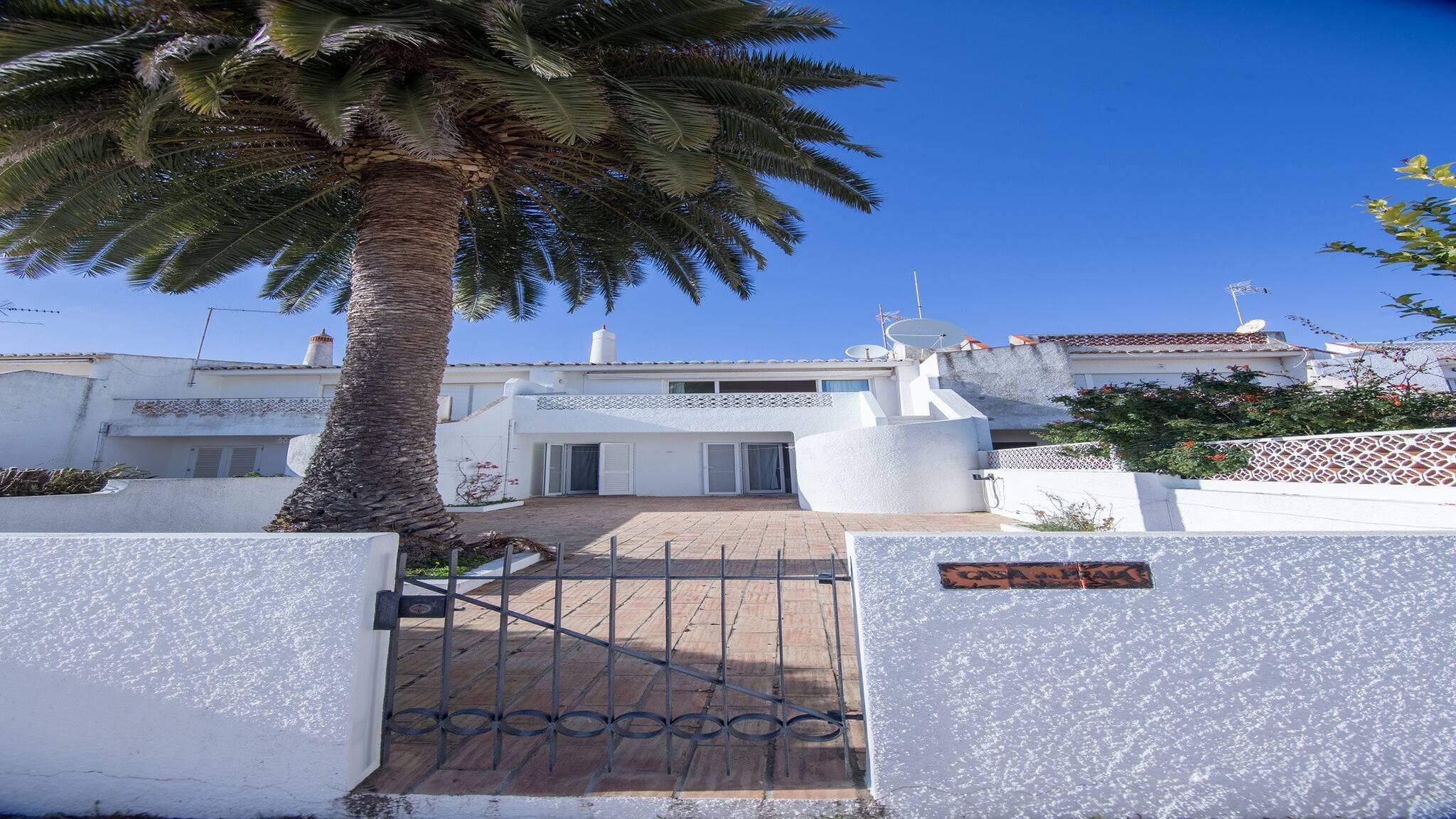 A gated entrance with a palm tree, a white building with a satellite dish, and a clear blue sky.