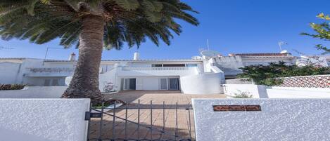 A gated entrance with a palm tree, a white building with a satellite dish, and a clear blue sky.