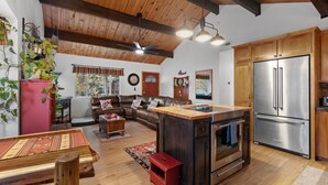 A spacious living area with a wooden ceiling, a large brown leather sofa, a dining table, and a kitchen with wooden cabinets and a stainless steel refrigerator. - Gorgeous cabin great for Family & Pets BBQ fenced backyard fire pit Jacuuzi (Big Bear City)