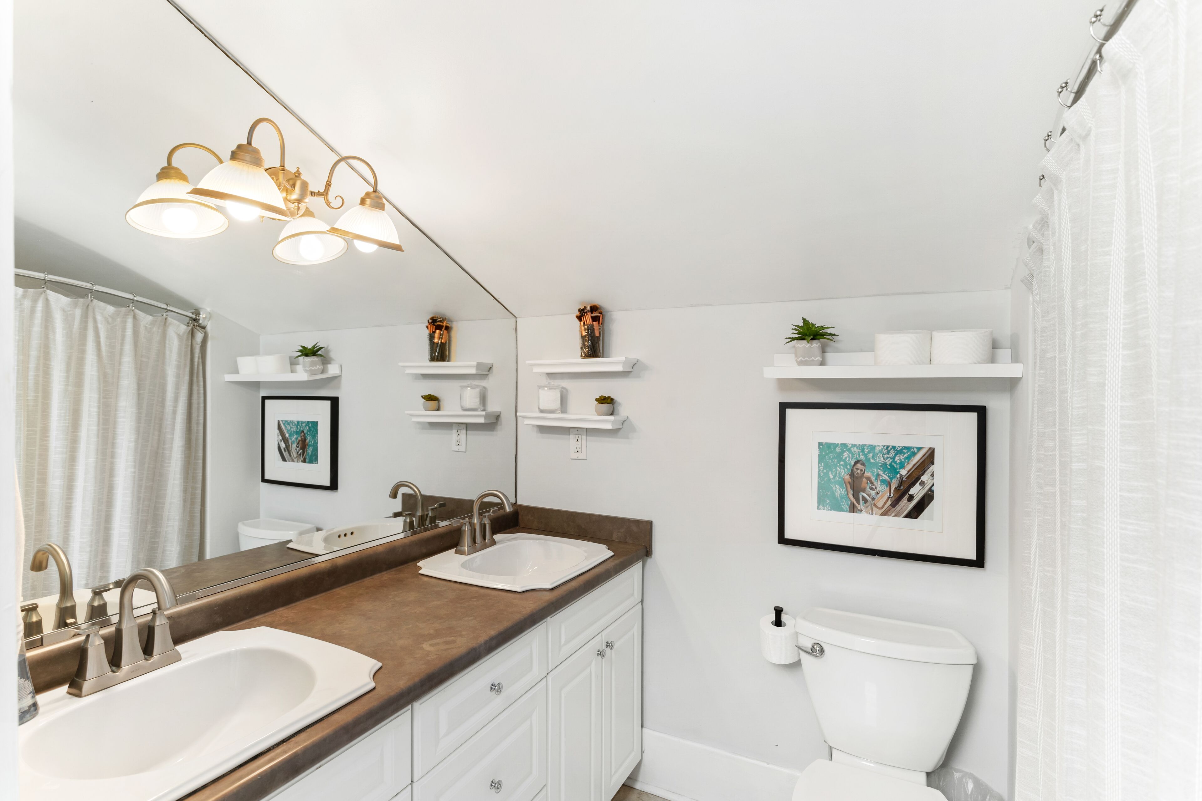 A modern bathroom with a double sink, a toilet, and a shower curtain. There are shelves with toiletries and a mirror above the sink.