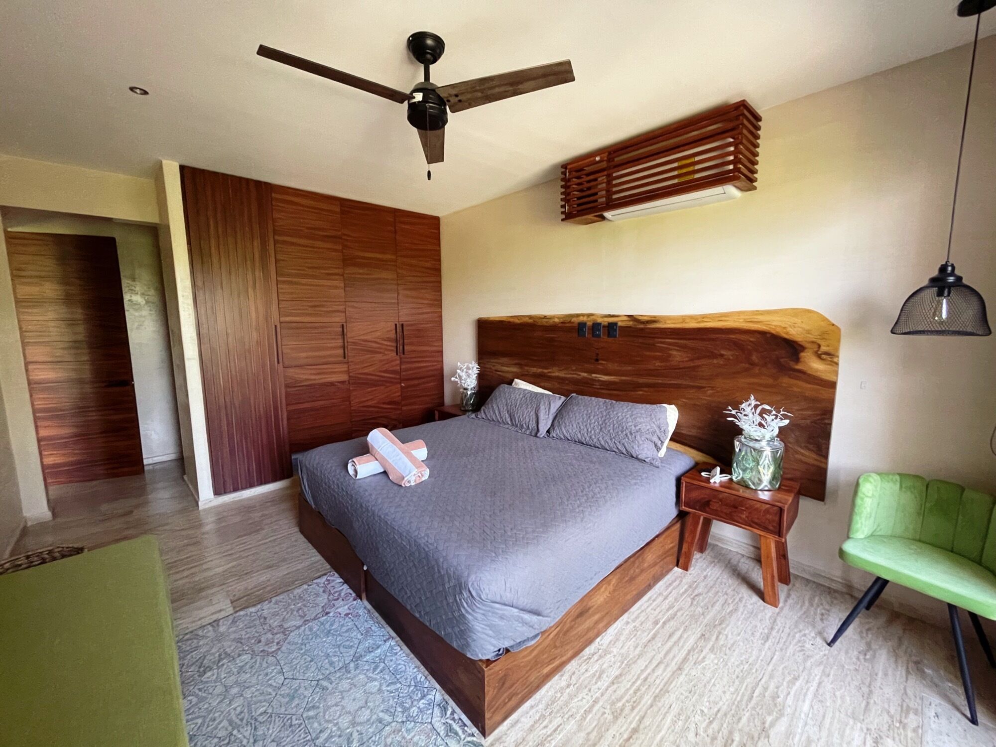 A bedroom with a large bed, wooden wardrobe, ceiling fan, bedside table, and a green chair.