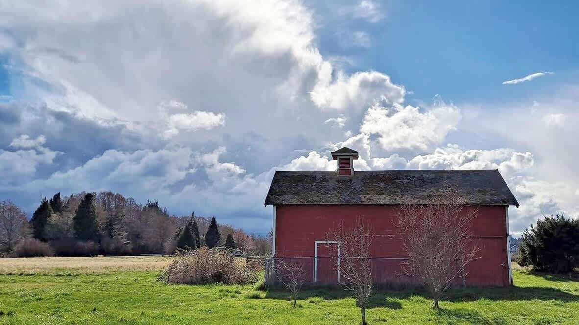 Un granero rojo con chimenea, rodeado de campos verdes y árboles bajo un cielo parcialmente nublado.