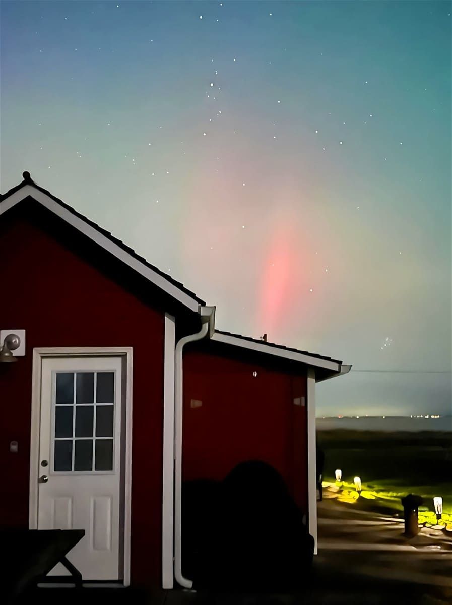 A red house with a white door and windows, under a night sky with visible stars and a faint aurora.