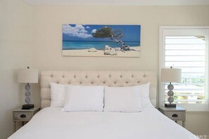 A bedroom with a large bed, two bedside tables, a painting of a beach scene, and a window with blinds. - Luxurious beach home with a beautiful ocean views. (Dana Point)