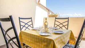 Une table dressée pour deux personnes, recouverte d’une nappe jaune, avec deux chaises et une bouteille de vin, sur un balcon offrant une vue panoramique.