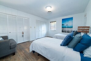 Quarto com cama, poltrona cinza, janela com persianas, luminária no teto e um quadro na parede.